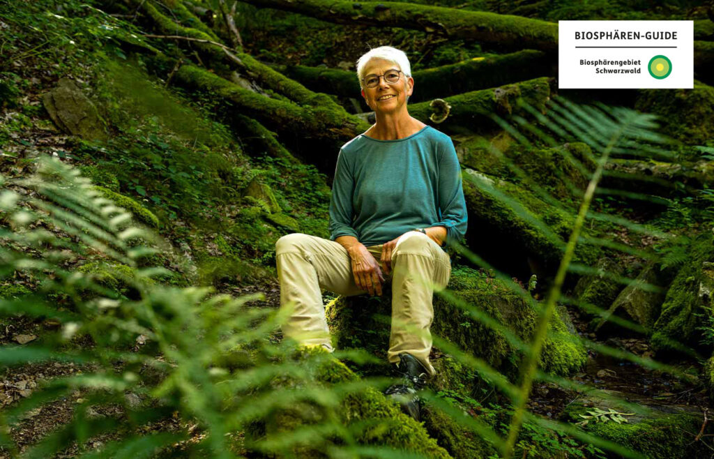 Ein Tour Guide sitzt im Wald auf Moos, davor sind Pflanzen